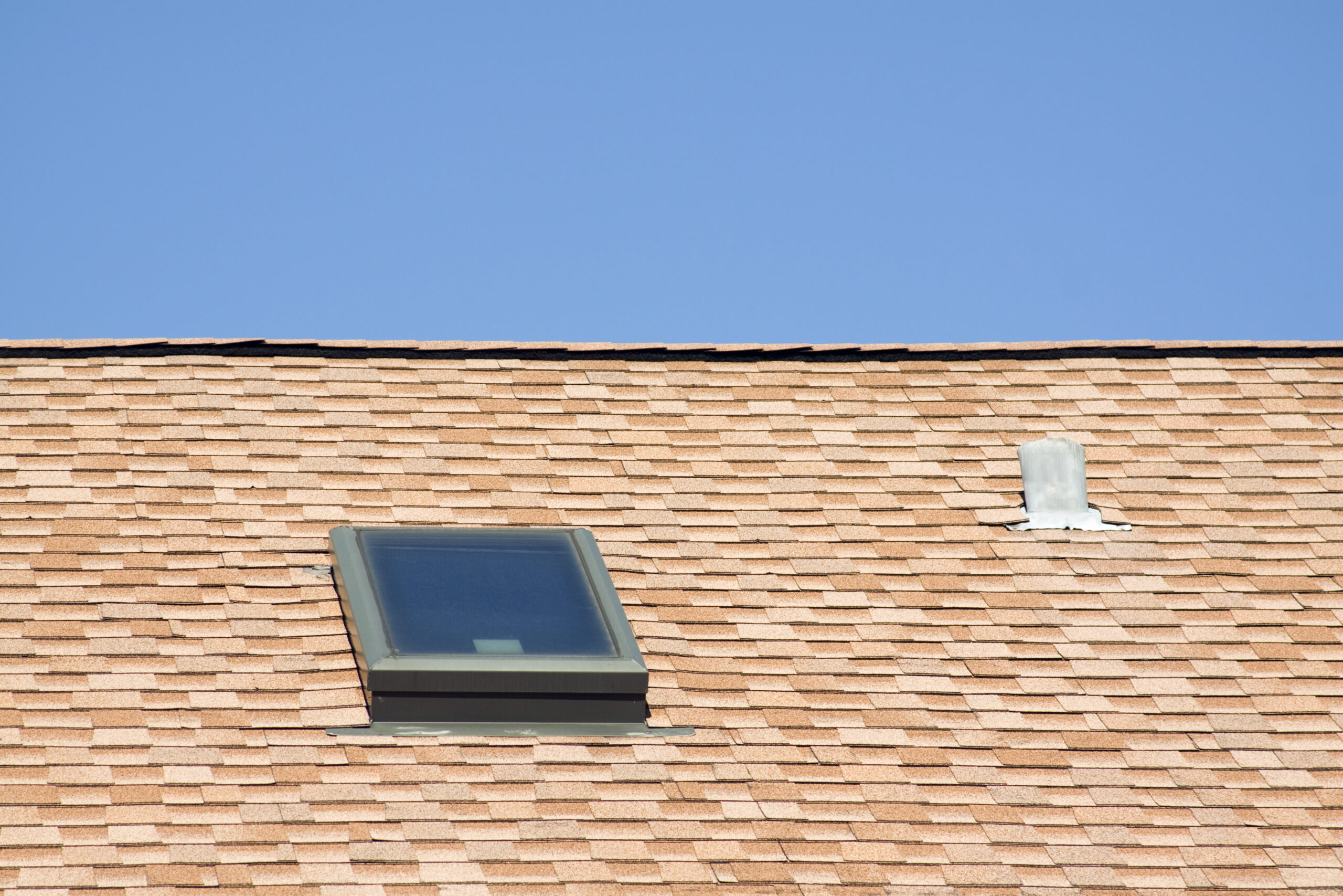 Close-up of a tan asphalt shingle roof featuring a rectangular skylight and a roof vent beneath a clear blue sky.